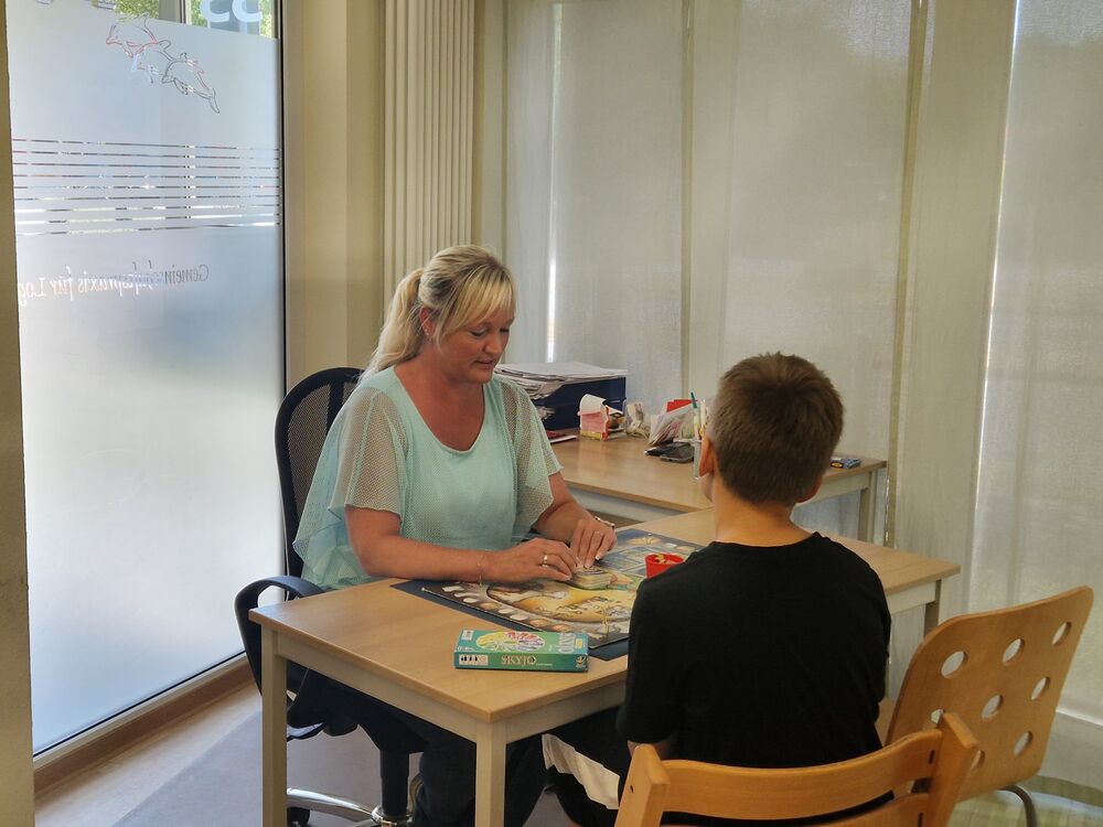 Denise arbeitet mit einem Kind an einem Tisch mit einem Spiel; das Kind ist von hinten zu sehen